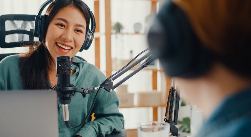 Une femme avec un casque d’écoute et un microphone sourit à une personne assise en face d’elle.